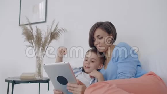 母亲和儿子在家里的卧室里使用数字平板电脑用数字显示快乐白人母子的正面观点视频的预览图