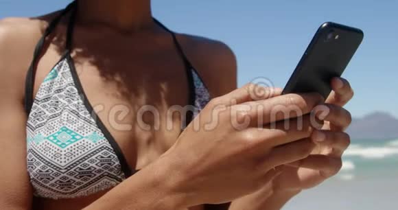 穿比基尼的年轻非洲裔美国妇女在海滩上使用手机的正面景色视频的预览图