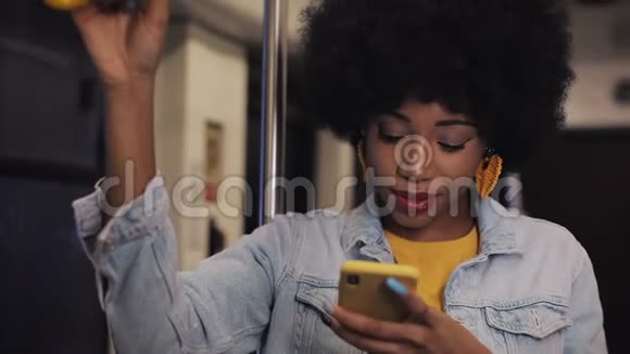微笑着年轻的非裔美国妇女握着扶手在公共交通工具上使用智能手机晚上雨水和雨水视频的预览图
