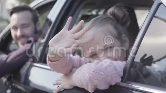 坐在汽车后座上的小女孩挥动着她的手近距离地看着镜头父亲在开车视频的预览图