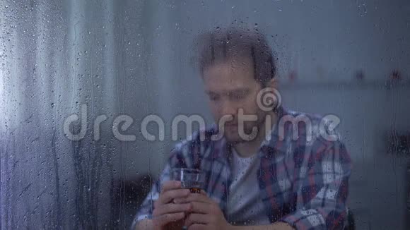 沮丧的男人在雨天独自喝威士忌想着生活中的问题视频的预览图