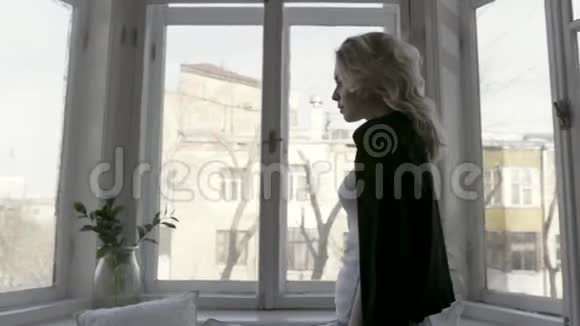 窗户边一个漂亮苗条的金发美女在家的侧景行动穿着黑色羊毛衫的年轻女孩视频的预览图