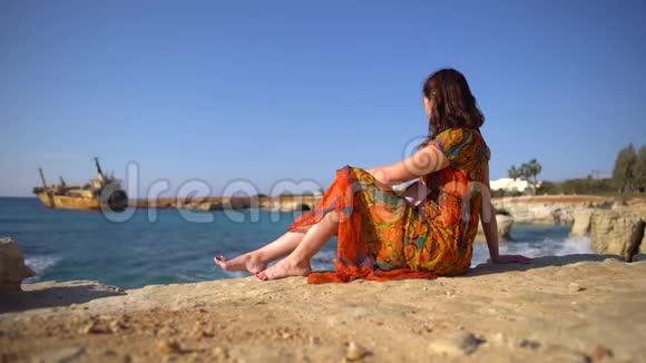 美丽的年轻女子穿着一件连衣裙坐在岩石海岸边背景是一艘古老的船天空和风景如画的景色视频的预览图