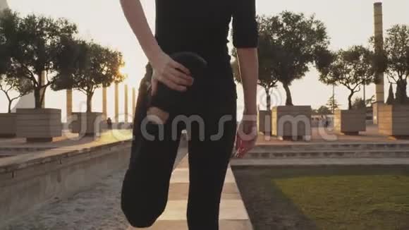 年轻女子穿着黑色运动服伸展腿部肌肉然后在阳光明媚的温暖天气里沿着街道慢跑少女视频的预览图