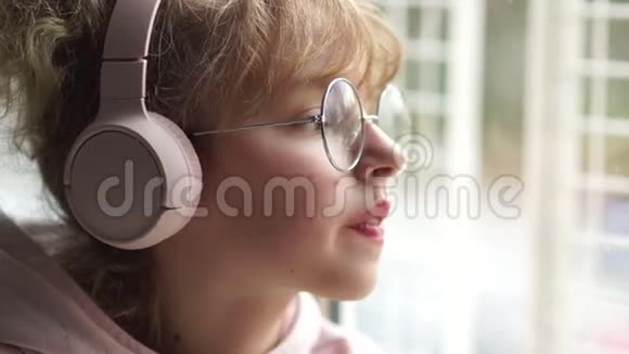 女学生听音乐很伤心窗户边一个戴着粉色耳机眼镜的女孩的肖像视频的预览图