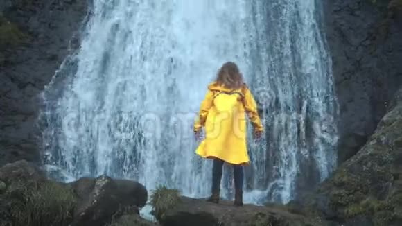 年轻的女旅行者徒步旅行者穿着黄色雨衣走到高地的瀑布前举起双手享受大自然视频的预览图