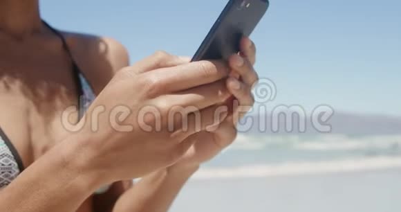 穿比基尼的年轻非洲裔美国妇女在海滩上使用手机的正面景色视频的预览图