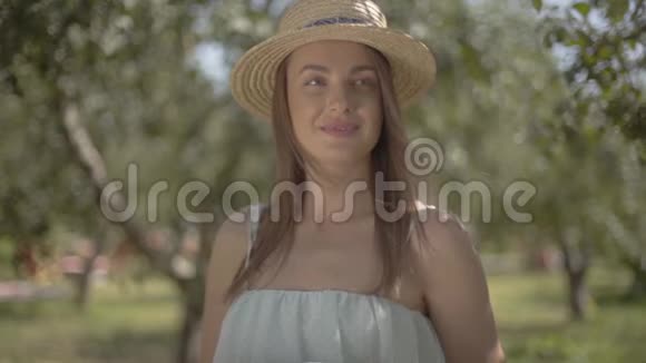 肖像可爱的年轻女子戴着草帽穿着白色长裙看着镜头微笑地站在绿色的夏天视频的预览图
