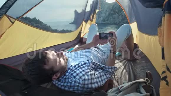 露营者躺在帐篷里用智能手机拍照脚一个年轻人躺在野营帐篷里在一张照片上视频的预览图