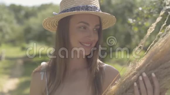可爱的年轻女子戴着草帽穿着长长的白色连衣裙站在绿色的夏日花园里手里拿着一堆干草农村视频的预览图