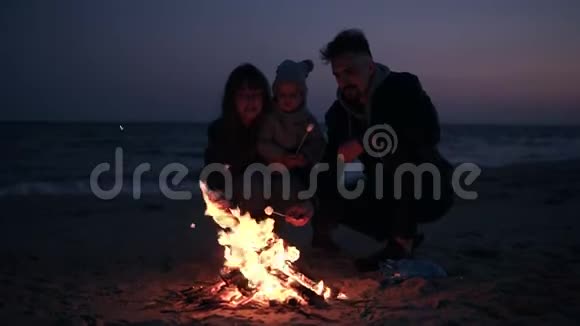 妈妈爸爸穿着暖和的衣服正在用木棍和他们的小宝宝烤棉花糖海滩上的篝火视频的预览图