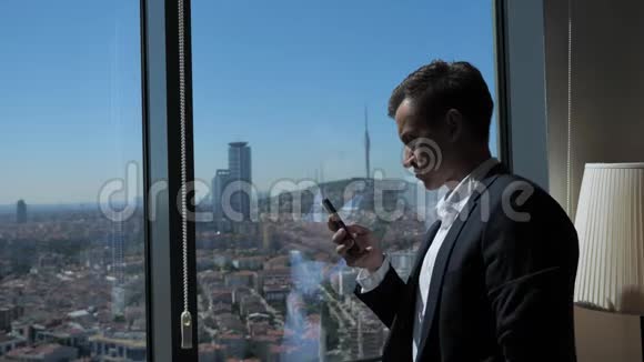 身着西装的年轻企业家正在窗口附近浏览电话可以看到全景城市视频的预览图
