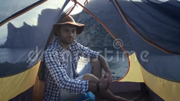 一个年轻人坐在野营帐篷里欣赏美丽的海景和山景徒步探险视频的预览图