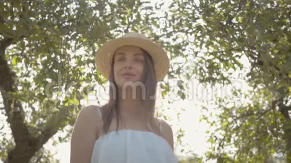 漂亮的年轻女子戴着草帽穿着长长的白色连衣裙站在绿色的里太阳在背后闪耀视频的预览图