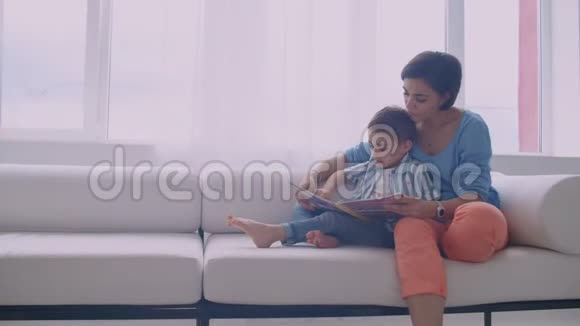 快乐的妈妈和小儿子早上一起在家客厅看书家庭活动概念视频的预览图