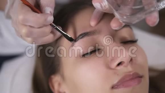 美学家正在用棕色染料在年轻女子的眉毛上涂上油漆在一家美容工作室里进行着色特写视频的预览图