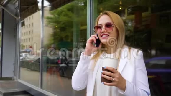 年轻时尚的金发女郎拿着咖啡杯在街上打电话视频的预览图