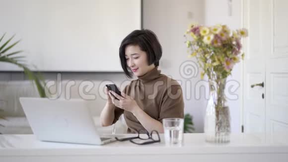 亚洲女人坐在白色的家庭办公室里滚动电话视频的预览图