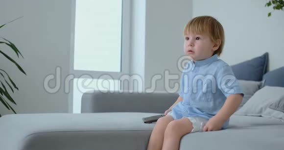 一个2岁的男孩坐在沙发上看电视手里拿着遥控器视频的预览图