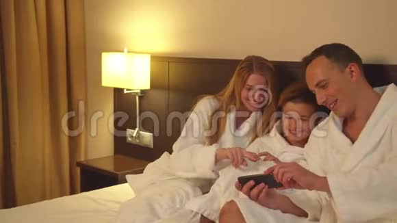 父母和儿子坐在床上在酒店房间里看电话照片视频的预览图