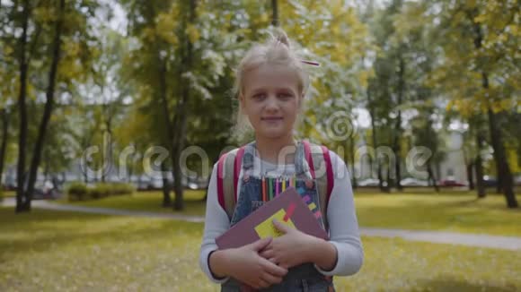 快乐的女学生带着背包拿着一本书住在学校院子里一个漂亮聪明的女孩微笑的肖像视频的预览图