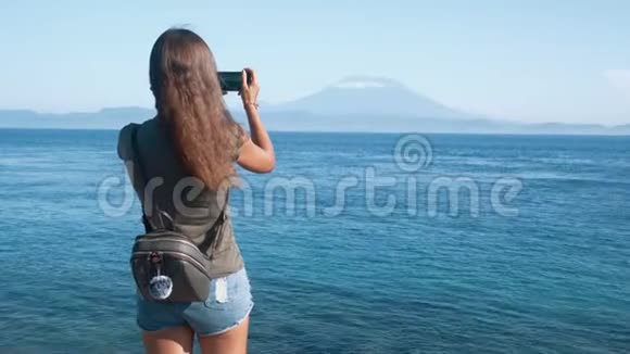 带背包的女孩旅行者的背面照片在电话中拍摄海洋和火山视频的预览图