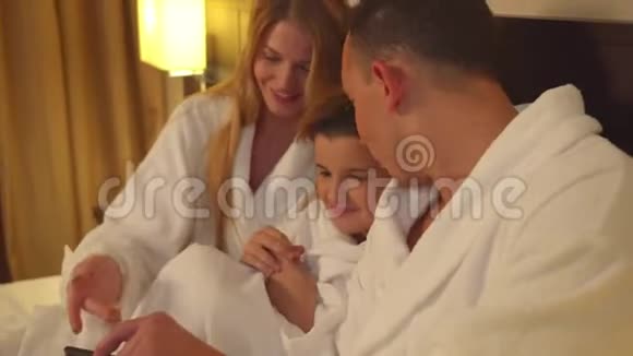 父母和儿子坐在床上在酒店房间里看电话照片视频的预览图