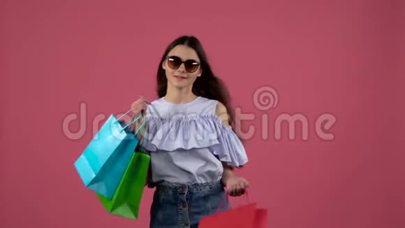 穿着太阳镜的女孩在购物后跳舞带着五颜六色的包裹粉色背景慢动作视频的预览图