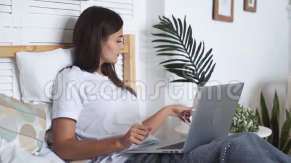 女孩坐在床上用笔记本电脑工作喝茶慢动作视频的预览图