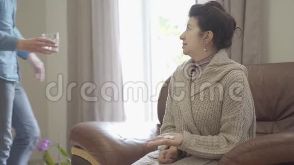这位成熟的女士坐在家里的皮椅上她病了成年的孙子带着水杯视频的预览图