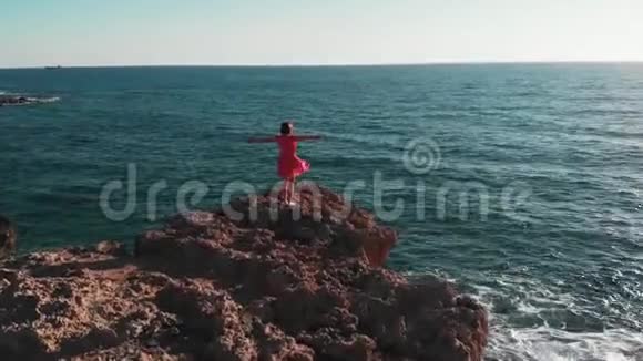 站在岩石峭壁上双手叉开对着太阳和太阳路的妇女剪影穿红衣服的女孩随风飘动视频的预览图