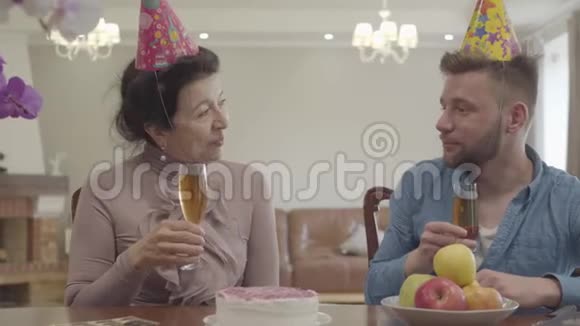 可爱的奶奶和成年孙子碰着坐在桌子旁的眼镜头上戴着生日帽成熟的女人视频的预览图