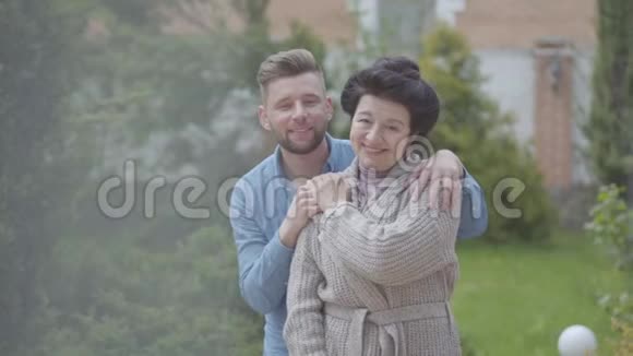 肖像可爱的成熟女人站在花园的大房子前面成年孙子拥抱她把她放在花园里视频的预览图
