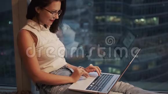 一个年轻的女人用一台笔记本电脑舒适地坐在高处的窗边视频的预览图