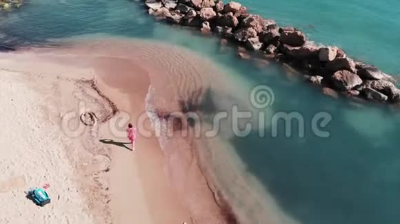 海滩上穿红色裙子的漂亮女人年轻女孩沿着海滩散步带蓝色清水的美丽沙滩鸟瞰视频的预览图
