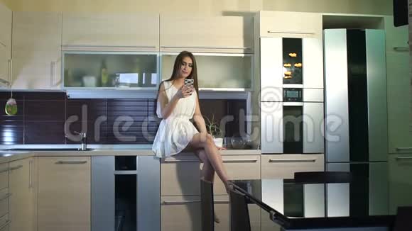 年轻的微笑女孩坐在厨房的台面上手里拿着电话视频的预览图