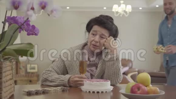 成熟的女士坐在桌子前的小蛋糕和苹果在盘子里成年孙子带来王冠和视频的预览图