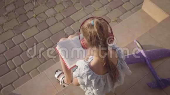 可爱的小女孩戴着耳机看书躺在门廊上的小飞机孩子花时间视频的预览图