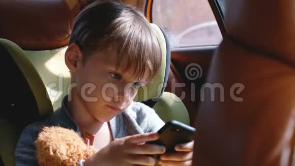 阳光明媚的一天4岁至6岁快乐的白人男孩在儿童安全座椅上使用智能手机带着玩具视频的预览图