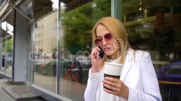 美丽的金发女人在手机上说话拿着咖啡杯在街上视频的预览图