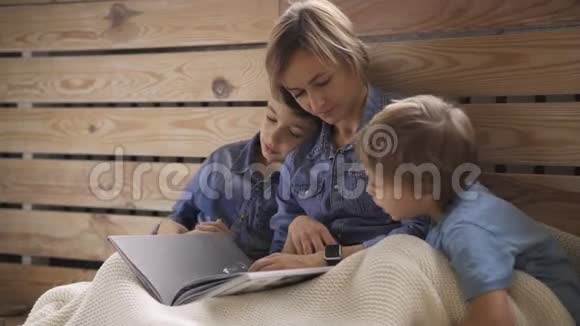年轻的白种人女人读一本书给两个坐在地板上的儿子一位美丽的年轻母亲正在看书视频的预览图