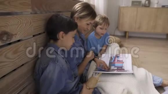年轻的白种人女人读一本书给两个坐在地板上的儿子一位美丽的年轻母亲正在看书视频的预览图