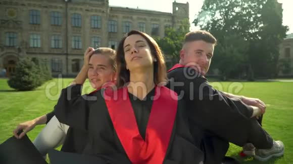 两个快乐的女孩和一个坐在壁炉里的男孩坐在大学大楼前的草地上年轻人庆祝视频的预览图