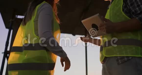 建筑工人拿着一块石碑一个戴着白盔的女检查员站在屋顶上在日落时分握手视频的预览图