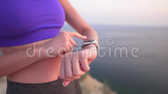 年轻的运动女孩在海边的悬崖上使用可穿戴的智能手表晚上或早晨触摸并滚动视频的预览图