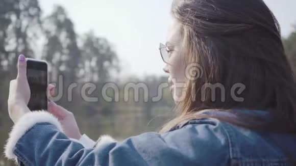 穿着牛仔裤夹克和眼镜在手机上拍照的漂亮迷人女人的后景视频的预览图