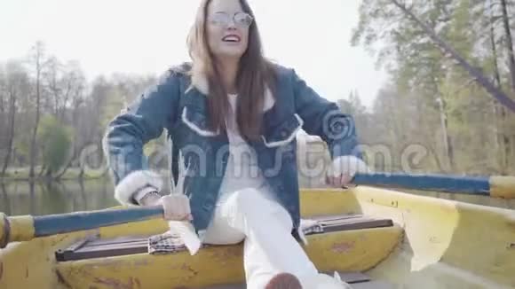 美丽的微笑魅力女人穿着白色裤子牛仔裤夹克和太阳镜在河上的黄船上划桨视频的预览图