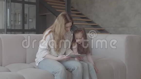 肖像画上漂亮的年轻母亲和她可爱的小女儿正在用平板电脑微笑着坐在大沙发上视频的预览图