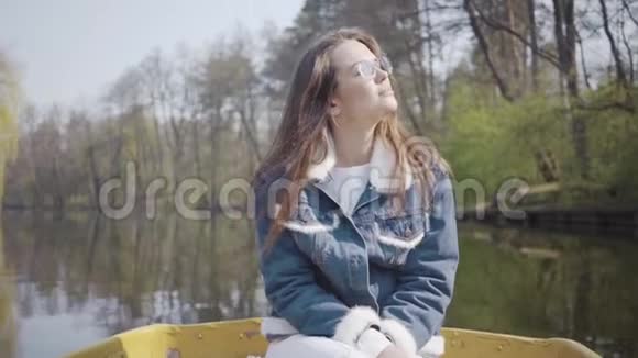 一个优雅的年轻女人戴着太阳镜和一件牛仔夹克漂浮在湖面或河流上的船上的肖像美丽美丽视频的预览图