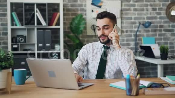 微笑的家伙在手机上说话在现代办公室里打笔记本电脑视频的预览图